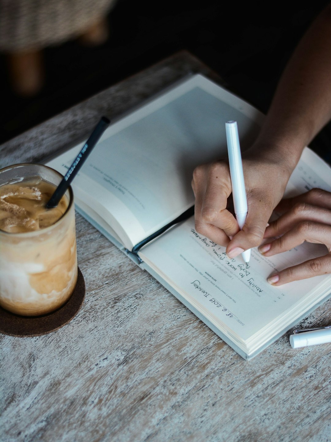 Mains écrivant dans un carnet avec un marqueur blanc et un café glacé.